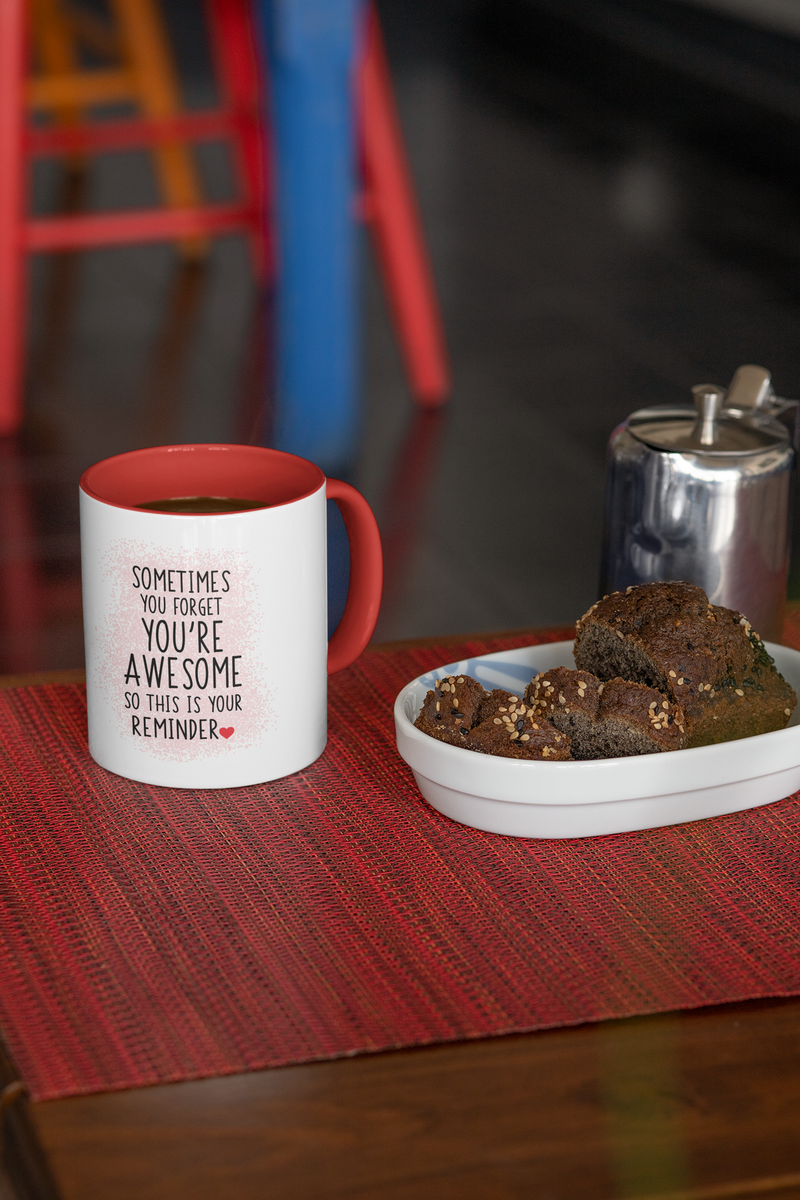 “Sometimes you forget you're awesome” coffee mug with a red handle, placed on a table next to brownies and a coffee pot.