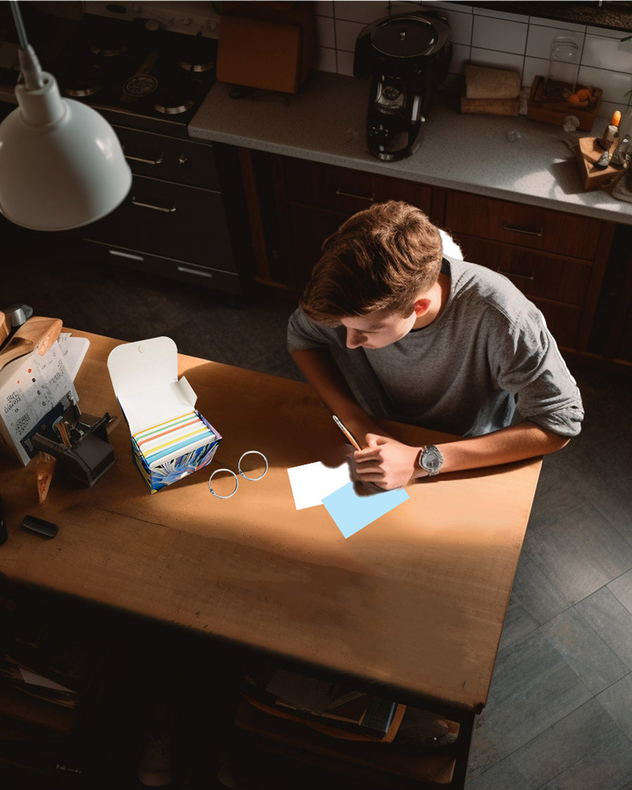 A person writing on a blue index card at a wooden table, with a box of multi-color blank index cards nearby.
