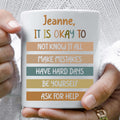 White ceramic mug with 'It Is Okay' reassurance quote held in hands.
