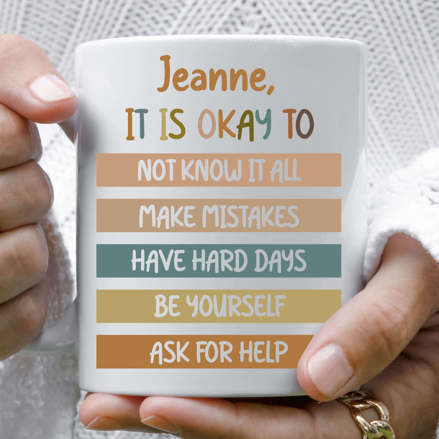 White ceramic mug with 'It Is Okay' reassurance quote held in hands.