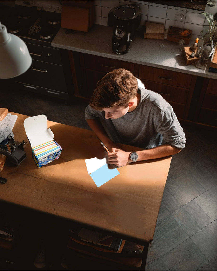 A person writing on a blue blank index card, with a box of multi-color index cards on a wooden table.
